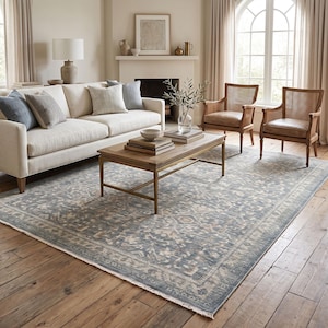 May include: A large rectangular area rug featuring a faded blue and beige floral pattern with a distressed finish and short fringe edges. The rug has a detailed central medallion and a wide decorative border, covering a wooden floor surface.