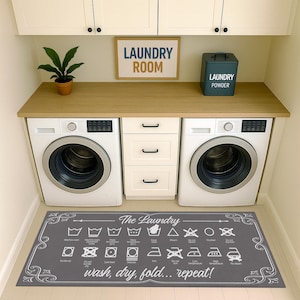 May include: A laundry room scene featuring two white washing machines, a wooden countertop, and a gray rug with laundry symbols and the text "wash, dry, fold... repeat!". A sign reads "LAUNDRY ROOM". A teal laundry powder container is on the counter.
