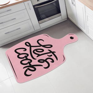 May include: A pink, cutting board-shaped kitchen mat with the words "Let's cook" in black script. The mat is placed on a tiled floor in front of a white kitchen cabinet and oven.