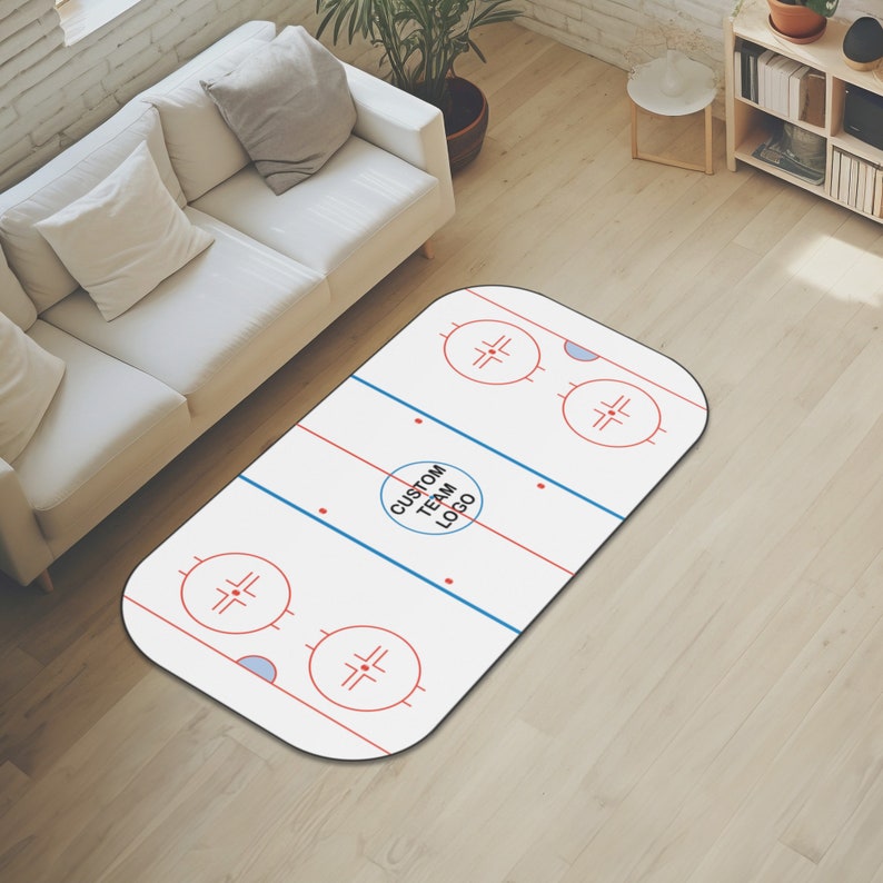 May include: White oval rug with a hockey rink design. The rug has red and blue lines, two blue circles with red crosses, and a circle in the center that says "Custom Team Logo".