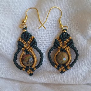 May include: A pair of green and gold macrame earrings with a round, speckled stone bead in the centre of each earring.