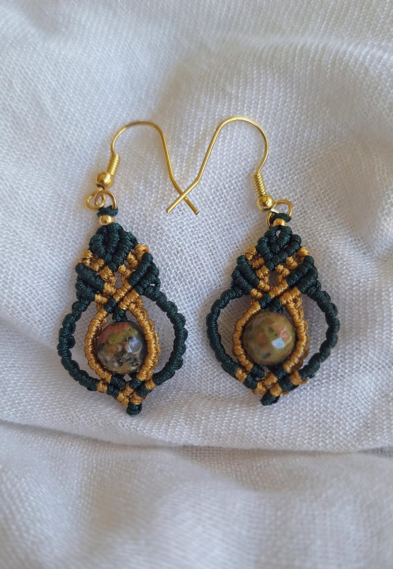 May include: A pair of green and gold macrame earrings with a round, speckled stone bead in the centre of each earring.