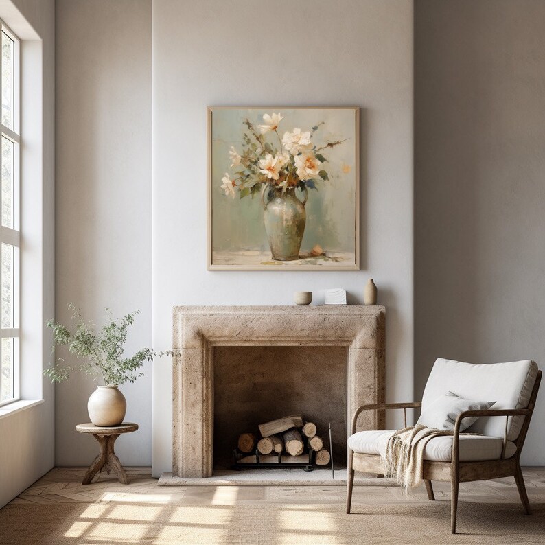 May include: A framed painting of a bouquet of white flowers in a green vase, hanging above a fireplace in a living room with a beige armchair and a small side table with a vase.