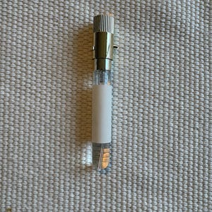 May include: A small, cylindrical glass perfume sample vial featuring a clear liquid interior. The vial is topped with a metallic silver cap that has a ribbed texture and includes a small side pin. A plain, rectangular white label wraps around the center.