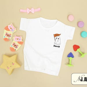 May include: A white t-shirt with a ghost graphic holding a flag that says "Boo" and the name "David". The image also includes a pink bow, socks with bear faces, a yellow star, and colorful toys.