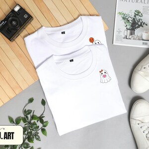 May include: Two white t-shirts with cartoon ghost designs. One shirt has a ghost holding a heart, the other has a ghost and a pumpkin. A black camera, white sneakers, and a magazine are also in the image. The brand name "NJ.ART" is visible.