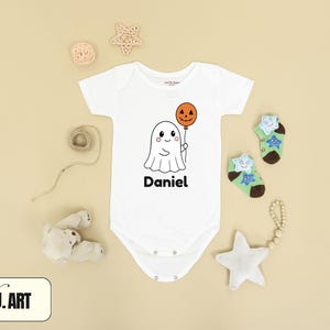 May include: White baby onesie with a cartoon ghost holding a jack-o'-lantern balloon. The name "Daniel" is printed below the ghost. The image is surrounded by Halloween-themed decorations, including star-shaped ornaments and socks.