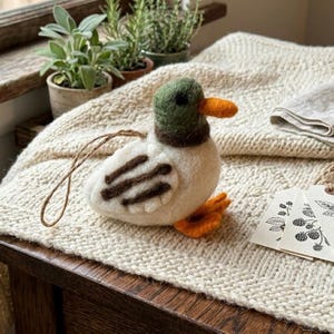 Peut inclure: Ornement de canard en feutre fait à la main. Le corps est blanc avec des détails bruns sur les ailes, un bec et des pieds orange, une tête verte et un cou brun. Une ficelle marron est attachée pour l'accrocher. L'ornement est sur une couverture en tricot crème.