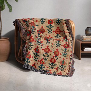 May include: A woven throw blanket featuring a repeating pattern of red and green floral motifs on a tan background. The blanket has a textured appearance with fringed edges along the sides and bottom, draped over a curved rattan chair.