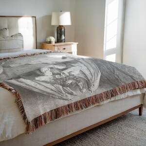 May include: A gray woven throw blanket with a detailed black and white illustration of a winged figure. The blanket has a fringed edge with brown, orange, and white threads. The blanket is draped over a bed.
