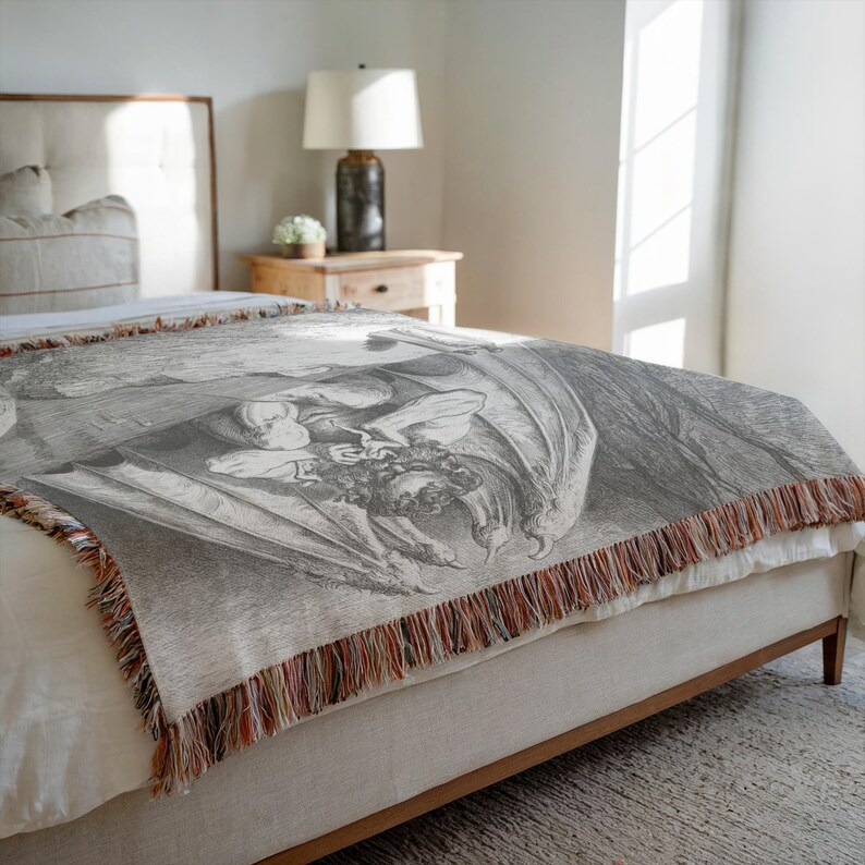 May include: A gray woven throw blanket with a detailed black and white illustration of a winged figure. The blanket has a fringed edge with brown, orange, and white threads. The blanket is draped over a bed.