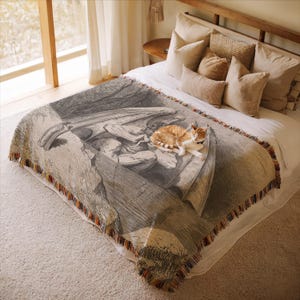 May include: A woven throw blanket with a detailed black and white illustration of a classical artwork, featuring a winged figure. The blanket has a fringed edge with red, orange, and brown accents. An orange and white cat is resting on the blanket.