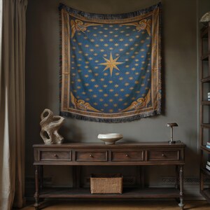 May include: A blue tapestry with a gold starburst and star pattern hangs on a wall. The tapestry has a fringe and angel figures. Below, a dark wood console table holds a white bowl, a woven basket, and a small lamp.