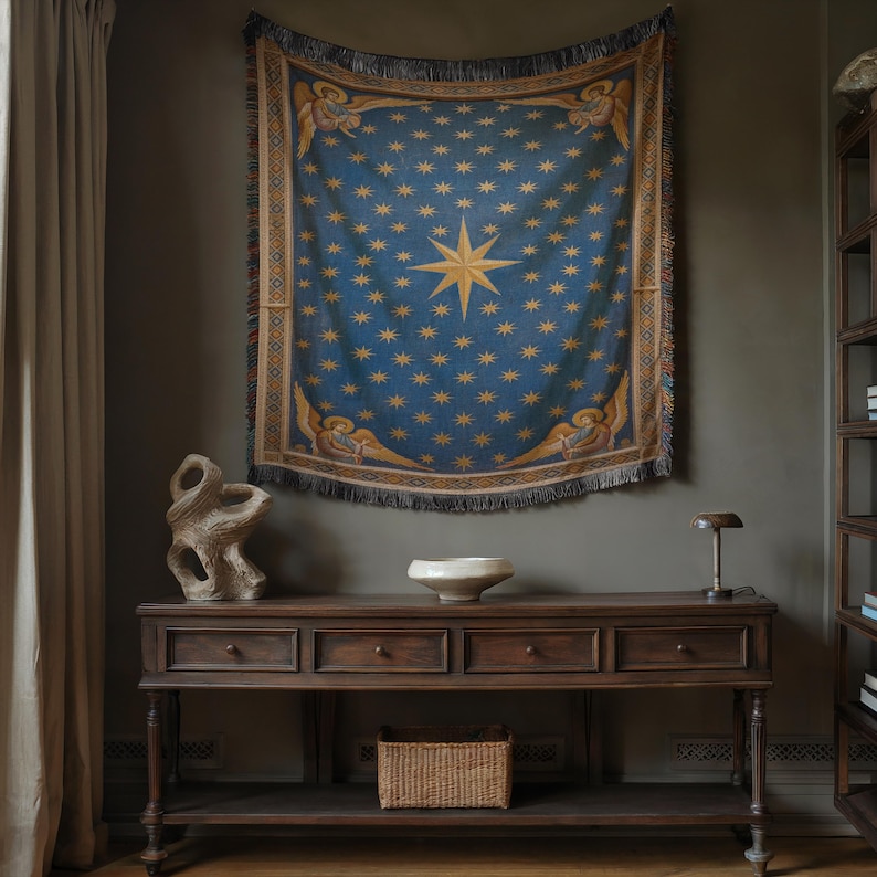 May include: A blue tapestry with a gold starburst and star pattern hangs on a wall. The tapestry has a fringe and angel figures. Below, a dark wood console table holds a white bowl, a woven basket, and a small lamp.