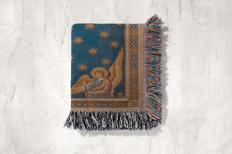 May include: A woven throw blanket with a celestial design. It has a blue background with gold stars and an angel with golden wings and a halo. The edges are trimmed with colourful fringe.