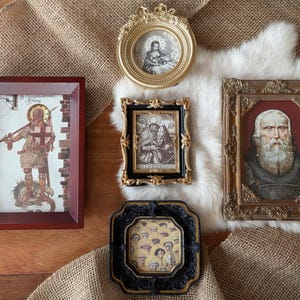 Peut inclure: Une sélection d'œuvres d'art encadrées, comprenant un portrait d'homme barbu, des icônes religieuses et d'autres illustrations vintage. Les cadres varient en style, de l'or orné au bois simple, et sont disposés sur une surface texturée.