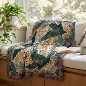 May include: A woven throw blanket featuring a repeating pattern of dark blue birds in flight amidst beige foliage, pine needles, and small red berries. The blanket has a fringed edge and is draped over a surface with an open book resting on top.