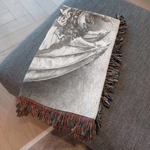 May include: A gray throw blanket with a detailed black and white illustration of a winged creature. The blanket has a fringe border with a mix of colorful threads. The blanket is folded over a gray ottoman.