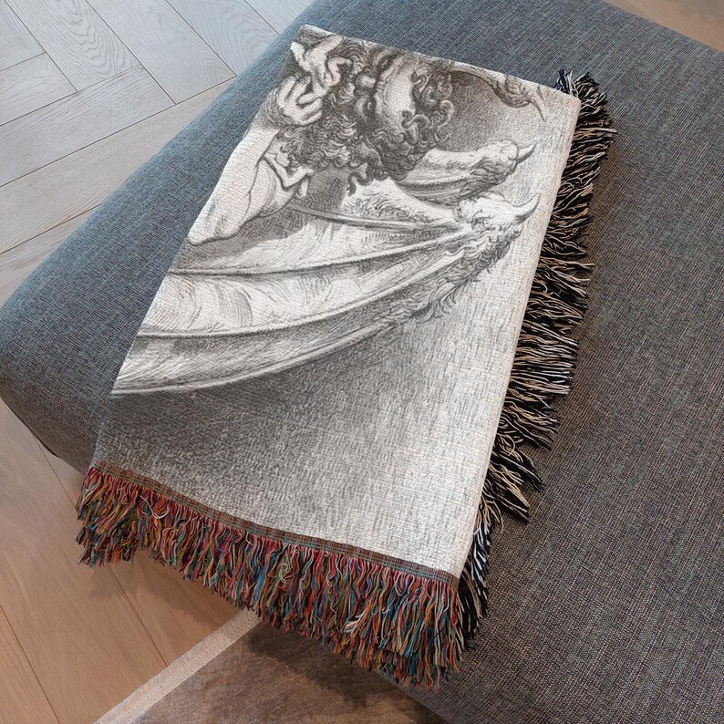 May include: A gray throw blanket with a detailed black and white illustration of a winged creature. The blanket has a fringe border with a mix of colorful threads. The blanket is folded over a gray ottoman.
