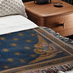 May include: A blue throw blanket with gold star and angel illustrations. The blanket has a fringed edge with multiple colours and is placed on a white bed. The blanket is likely made of woven fabric.