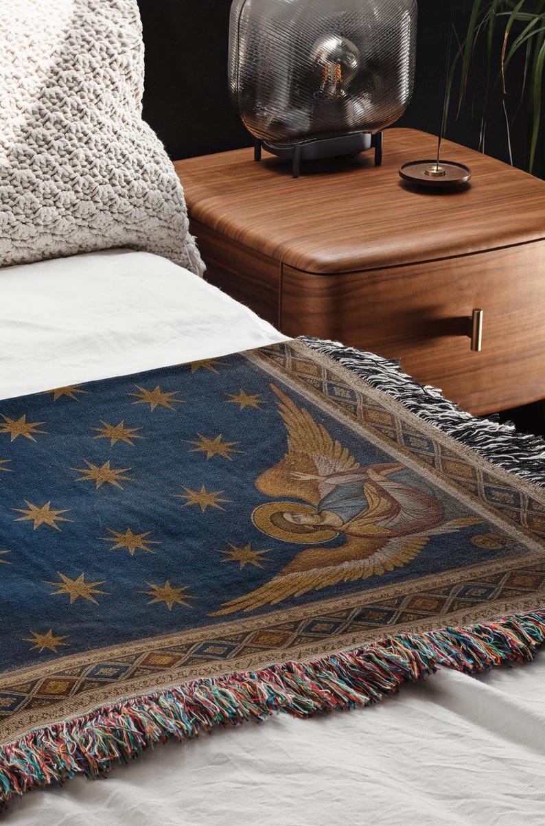 May include: A blue throw blanket with gold star and angel illustrations. The blanket has a fringed edge with multiple colours and is placed on a white bed. The blanket is likely made of woven fabric.