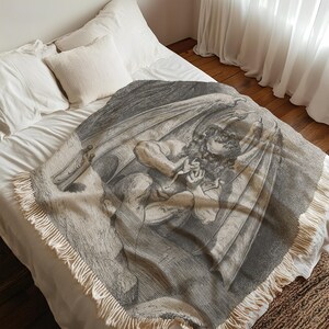 May include: A woven throw blanket featuring a black and white illustration of a winged figure. The blanket has a fringed edge and is draped over a bed with white bedding. The artwork depicts a classical scene.