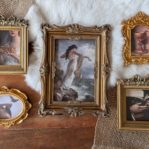 May include: A selection of framed art prints in an antique style. The frames are gold and ornate, featuring various classical paintings. The art includes a woman holding a box, a woman on a cliff, a chalice, a woman with a bird, and a person reading a book.