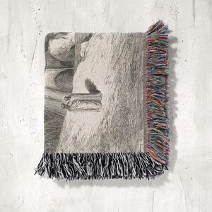 May include: A woven throw blanket with a black and white illustration of a figure in a room. The blanket has a fringe border with black, red, blue, and yellow threads. The blanket is displayed against a white textured wall.