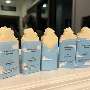 Pode incluir: Cinco barras de sabão com um topo fofo em forma de nuvem, embrulhadas em papel azul com gráficos de nuvens brancas. O texto "Head in the Clouds" e "Gooseneck Soap Company" estão impressos na embalagem.