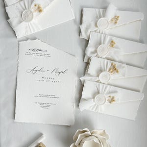 Op de afbeelding: Witte huwelijksuitnodigingen met een waxzegel en gedroogde bloemen. De uitnodiging luidt "In de naam van Allah, de meest barmhartige, de meest genadige. Angelina & Nayfal Maandag 14 april Samen met onze familie nodigen we u uit om onze bruiloft te vieren om 18:00 uur Daris Studio Marrakech, Marokko"