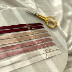 May include: A set of seven personalized ribbon tags with names written in gold lettering. The ribbons are in various shades of pink, beige, and white.
