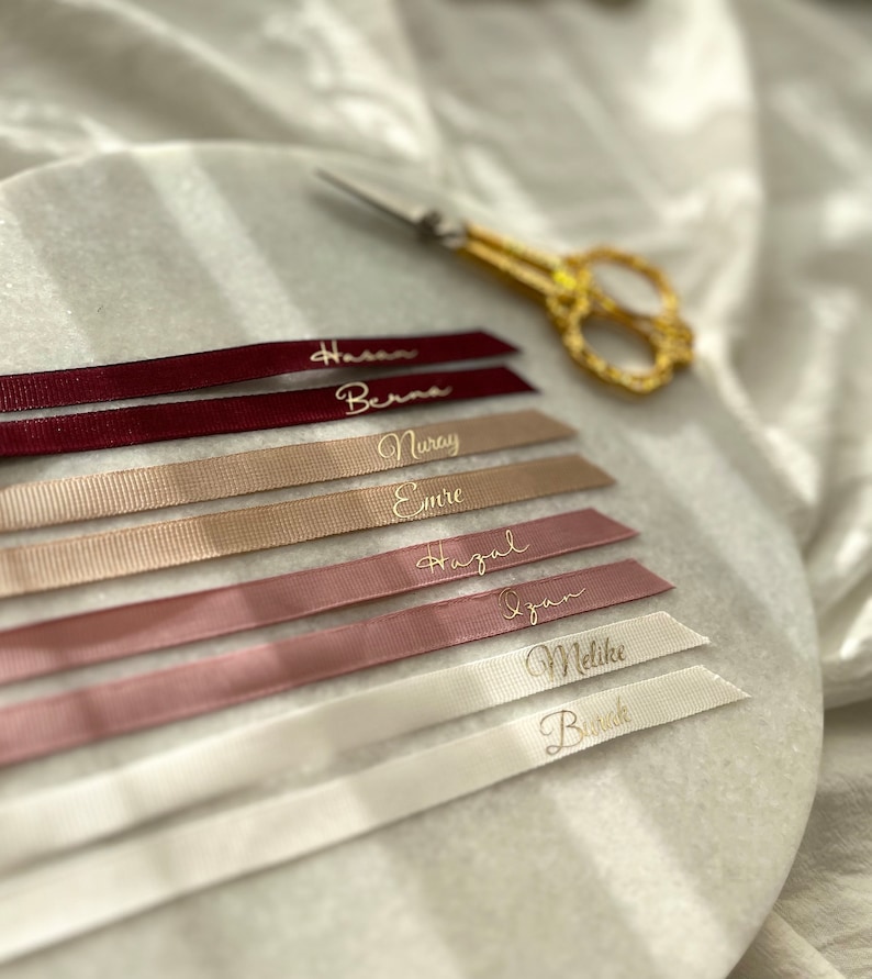 May include: A set of seven personalized ribbon tags with names written in gold lettering. The ribbons are in various shades of pink, beige, and white.