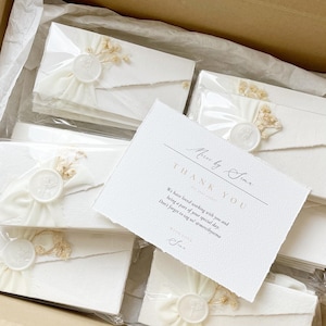Op de afbeelding: Een verzameling witte enveloppen met geschepte randen, elk verzegeld met een wit waszegel en versierd met gedroogde bloemen. Een witte bedankkaart met zwarte tekst Merci by Sema ligt bovenop de verpakte enveloppen.