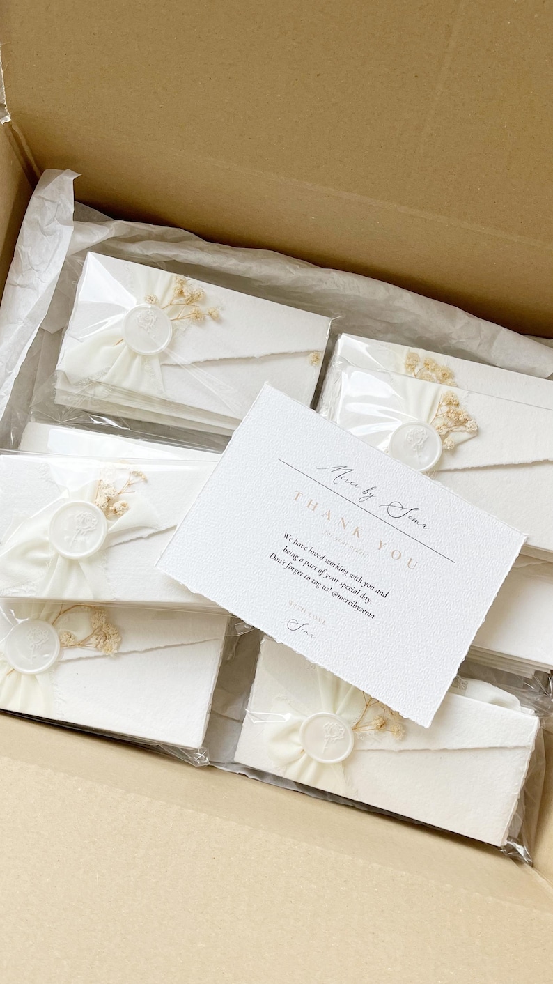 Op de afbeelding: Een verzameling witte enveloppen met geschepte randen, elk verzegeld met een wit waszegel en versierd met gedroogde bloemen. Een witte bedankkaart met zwarte tekst Merci by Sema ligt bovenop de verpakte enveloppen.