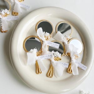 Gepersonaliseerde trouwbedankjes met kleine gouden handspiegeltjes, versierd met wit lint en droogbloemen, gepresenteerd op een keramisch bord – luxe en elegante wedding favors voor gasten.