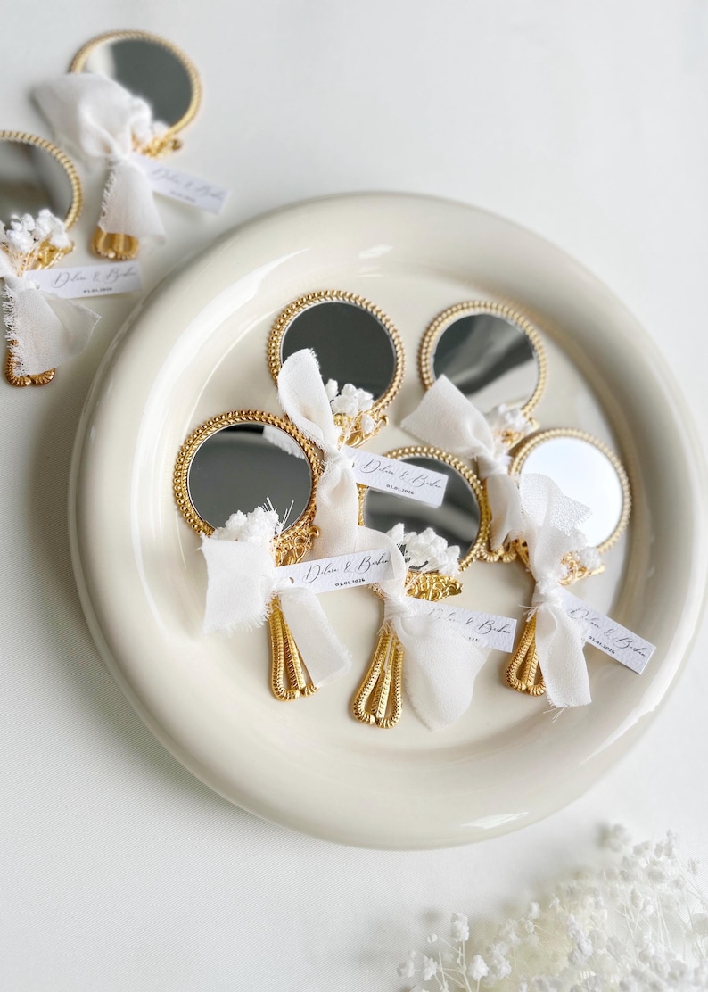 Gepersonaliseerde trouwbedankjes met kleine gouden handspiegeltjes, versierd met wit lint en droogbloemen, gepresenteerd op een keramisch bord – luxe en elegante wedding favors voor gasten.