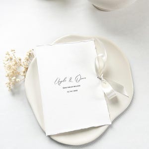 May include: A white invitation card with torn edges, tied with a satin ribbon, rests on a white plate. The card features elegant calligraphy with names and a date. Dried flowers are placed on the side, creating a minimalist wedding theme.
