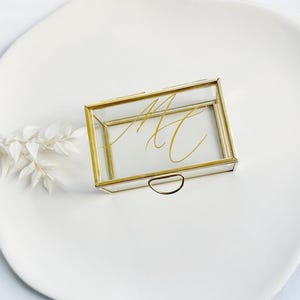 Op de afbeelding: Een rechthoekige glazen doos met gouden rand en een klein handvat. De doos bevat gouden kalligrafie initialen. De doos staat op een wit bord, met witte bloemaccenten.