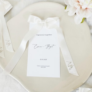 May include: A white invitation card with a white satin ribbon and bow. The card has the names "Esma - Yigit" and the date "25.10.2025" printed on it. The ribbon has the initials "Ey" printed on it. The card is on a white plate.