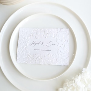 May include: A white wedding invitation with floral embossing, featuring the names "Hazal & Eren" and the text "welcome to our wedding." The invitation rests on a white plate, with a second plate beneath it. White floral accents are in the lower right corner.