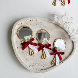 Gepersonaliseerde trouwbedankjes met kleine gouden handspiegeltjes, versierd met rood strikje en droogbloemen, gepresenteerd op een keramisch bord – luxe en elegante wedding favors voor gasten.