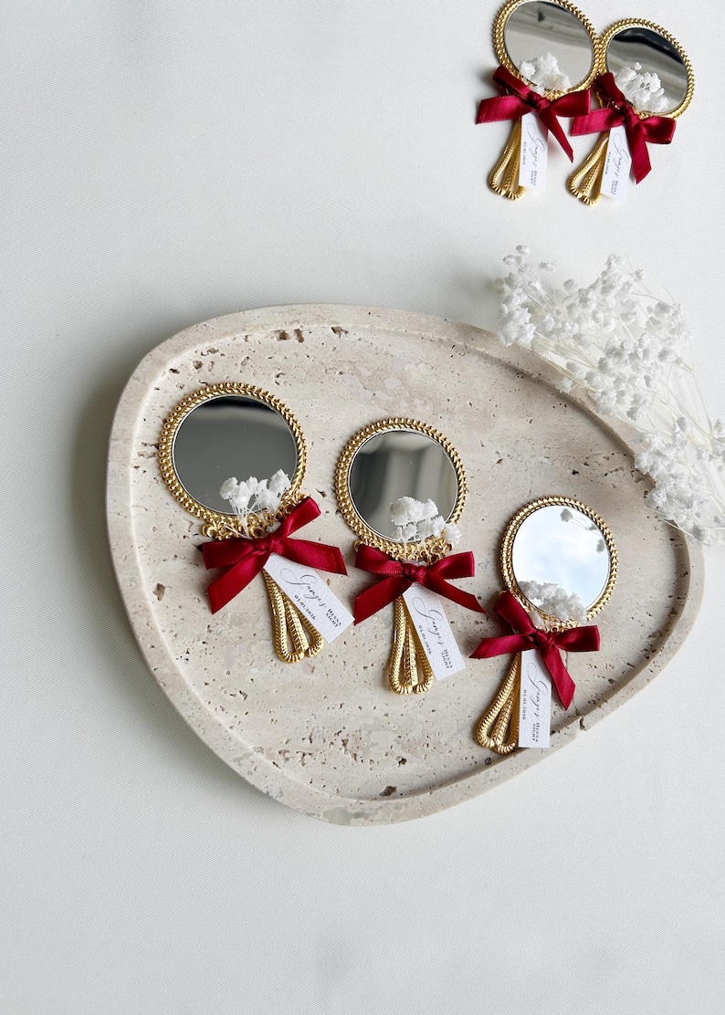 Gepersonaliseerde trouwbedankjes met kleine gouden handspiegeltjes, versierd met rood strikje en droogbloemen, gepresenteerd op een keramisch bord – luxe en elegante wedding favors voor gasten.