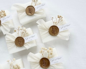 May include: Five white gift boxes wrapped in white tissue paper with a gold wax seal and a small sprig of dried white flowers. Each box has a white label with the text "Cyndi & Michael" and the date "01.01.2024".