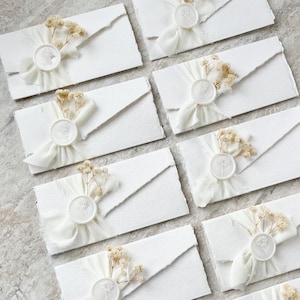 Op de afbeelding: Een verzameling witte rechthoekige enveloppen met gescheurde randen, elk verzegeld met een witte waszegel met een bloemmotief. Een klein takje gedroogde gipskruidbloemen en een wit chiffon lint zijn onder elke zegel bevestigd.