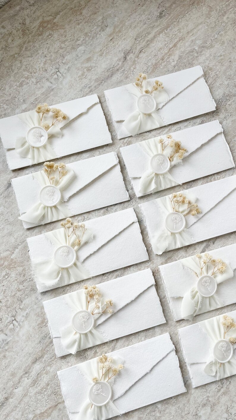 Op de afbeelding: Een verzameling witte rechthoekige enveloppen met gescheurde randen, elk verzegeld met een witte waszegel met een bloemmotief. Een klein takje gedroogde gipskruidbloemen en een wit chiffon lint zijn onder elke zegel bevestigd.