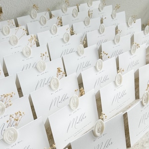 Op de afbeelding: Een verzameling witte placecards met een waszegel en gedroogde bloemen. De placecards zijn voor een huwelijksreceptie en hebben de namen "Mr. and Mrs. Mulder" erop gedrukt.