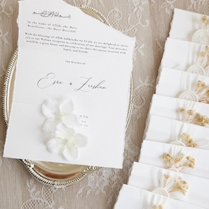 Op de afbeelding: Een witte trouwkaart van gestructureerd papier met gescheurde randen en zwarte bedrukte tekst, inclusief de namen Esra en Zeshan, versierd met twee witte stoffen bloemen. Een stapel bijpassende witte enveloppen met waszegels en gedroogde bloemen.