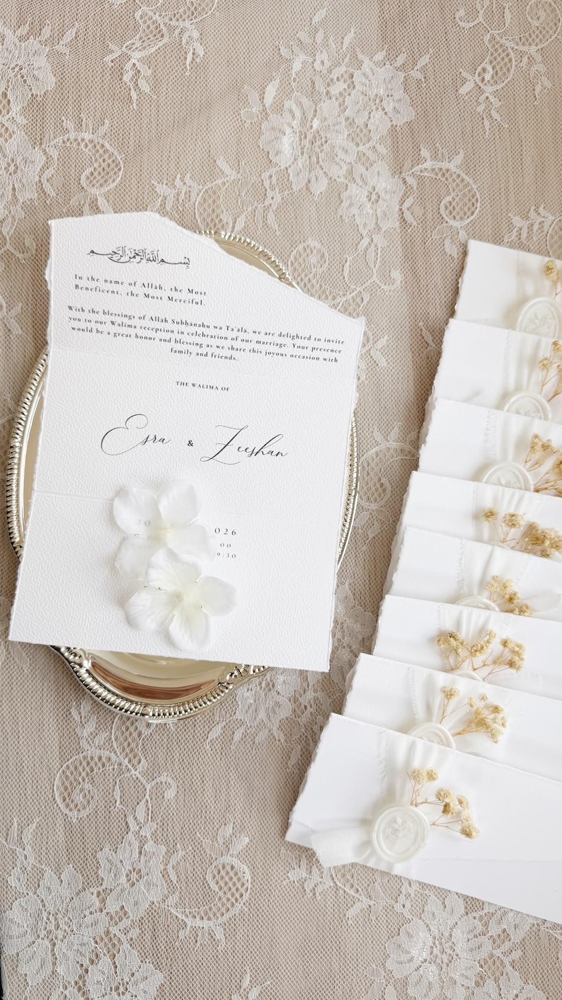 Op de afbeelding: Een witte trouwkaart van gestructureerd papier met gescheurde randen en zwarte bedrukte tekst, inclusief de namen Esra en Zeshan, versierd met twee witte stoffen bloemen. Een stapel bijpassende witte enveloppen met waszegels en gedroogde bloemen.
