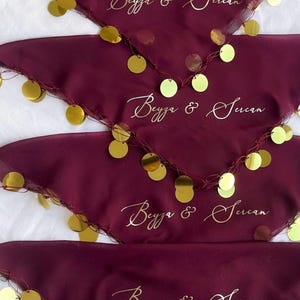 Puede incluir: Tela de color burdeos con adornos dorados y el texto "Beyza & Sercan" en escritura elegante. La tela está adornada con detalles dorados en forma de moneda a lo largo de los bordes. El diseño sugiere un artículo de celebración u ocasión especial.