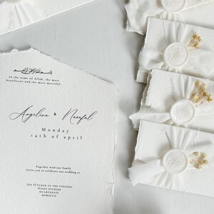 Op de afbeelding: Een witte trouwkaart met gescheurde randen en zwarte tekst met de namen Angelina en Naufal, samen met bijpassende witte enveloppen verzegeld met witte waszegels, wit lint en kleine gedroogde bloemen.
